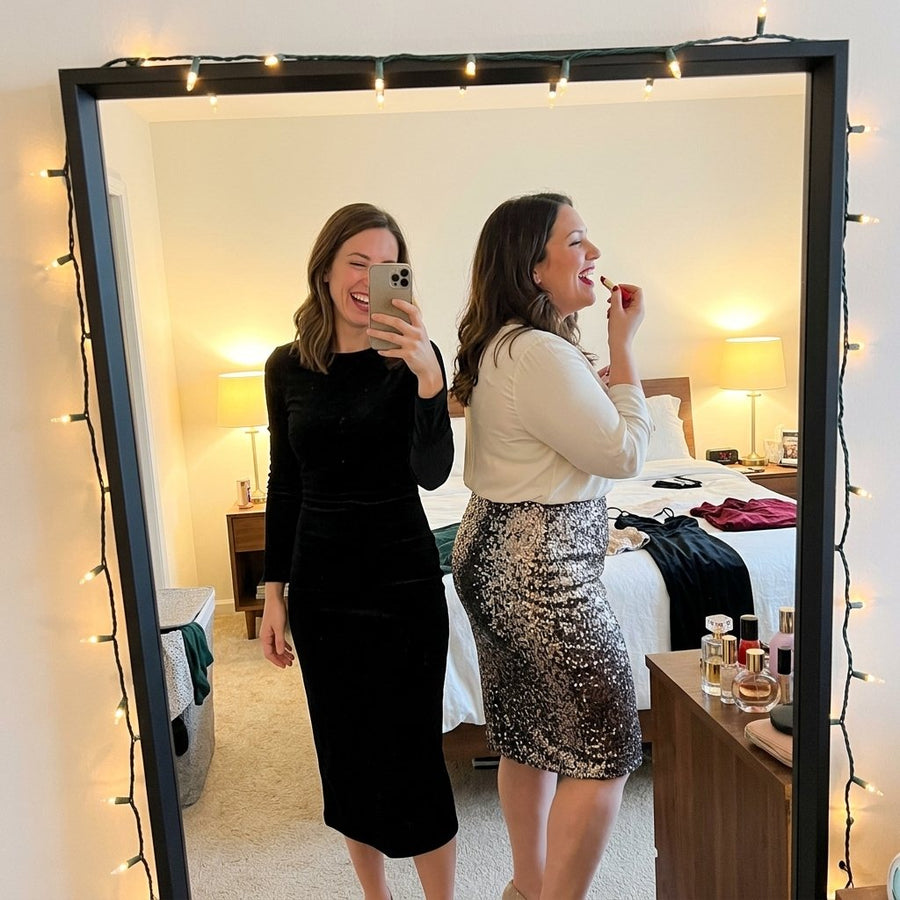 Two friends of different sizes laughing in a bedroom mirror selfie while getting ready for a holiday party